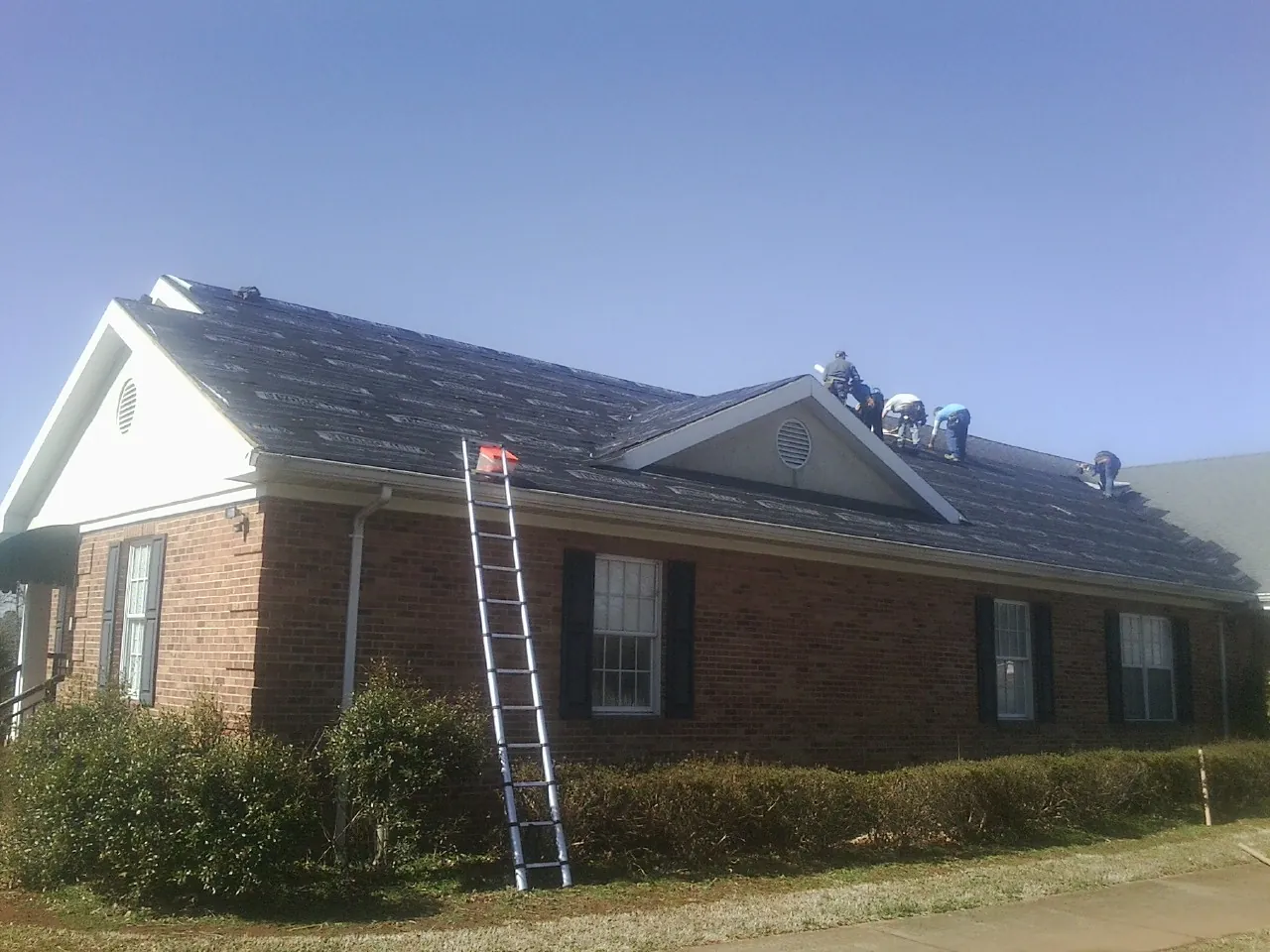 Crew tearing shingles off a brick ranch roof with a ladder set — Emergency Roof Repair work by our crew in Byron